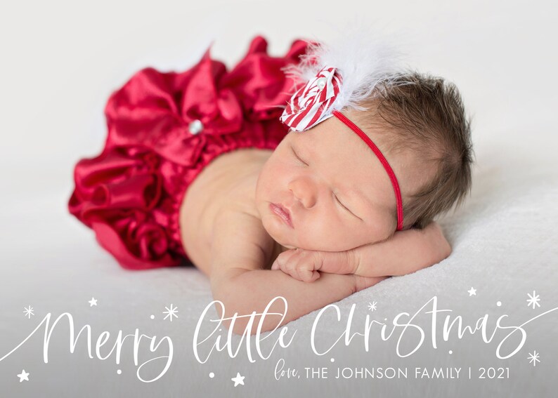 May include: A newborn baby girl wearing a red satin outfit with a white bow and a red and white candy cane bow on her head. The baby is sleeping with her head resting on her arm. The text "Merry little Christmas" is written in white cursive script below the baby. The text "love, THE JOHNSON FAMILY | 2021" is written in white below the cursive text.