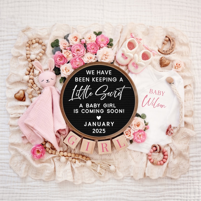 K&ouml;nnte beinhalten: Ein schwarzes Buchstabenbrett mit dem Text "We have been keeping a little secret. A baby girl is coming soon! January 2025." Das Brett ist umgeben von rosa und wei&szlig;en Blumen, einem rosa Baby-Outfit, einem Holzspielzeug und Holzbl&ouml;cken, die "GIRL" buchstabieren.