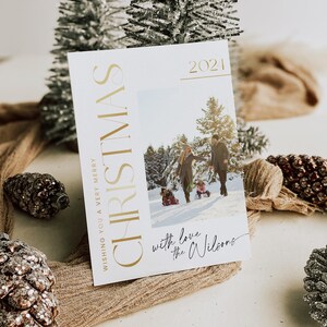 May include: A white Christmas card with gold lettering that reads "Wishing you a very merry Christmas 2021". The card features a photo of a family of four in a snowy forest. The text "with love the Wilsons" is written in black cursive at the bottom of the card.