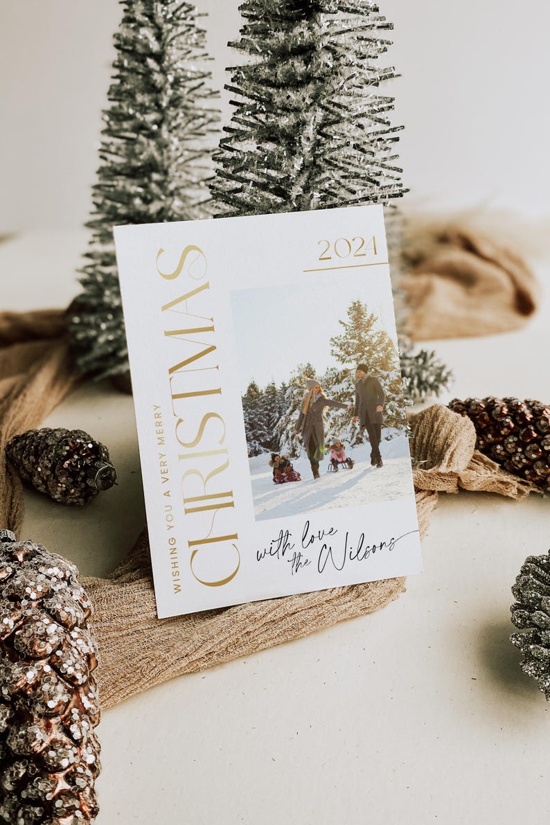 May include: A white Christmas card with gold lettering that reads "Wishing you a very merry Christmas 2021". The card features a photo of a family of four in a snowy forest. The text "with love the Wilsons" is written in black cursive at the bottom of the card.