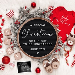 May include: A black letter board with the text "A Special Christmas Gift is Due to be Unwrapped June 2024" surrounded by red and white Christmas decorations, including a red baby onesie with the text "Baby Williams" and a crocheted reindeer toy.
