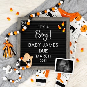 May include: A black letter board with the text "It's a Boy! Baby James Due March 2023". The board is surrounded by Halloween decorations, including a black and white ghost rattle, a black and white beaded garland, and a black and orange baby onesie with a ghost and pumpkin design.