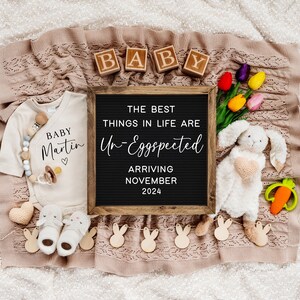 May include: A black letter board with the text "The best things in life are Un-Eggspected" and "Arriving November 2024".  A white onesie with the text "Baby Martin" and a heart.  A white bunny plush toy with a pink heart on its chest.  A wooden block spelling "BABY".  A carrot-shaped teether.  A garland of wooden bunny shapes.  A pair of baby shoes.  A heart-shaped crochet toy.  A bouquet of pink, yellow, and orange tulips.