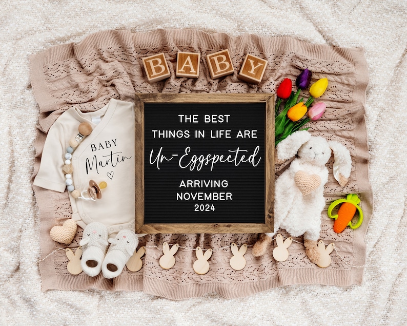 May include: A black letter board with the text "The best things in life are Un-Eggspected" and "Arriving November 2024".  A white onesie with the text "Baby Martin" and a heart.  A white bunny plush toy with a pink heart on its chest.  A wooden block spelling "BABY".  A carrot-shaped teether.  A garland of wooden bunny shapes.  A pair of baby shoes.  A heart-shaped crochet toy.  A bouquet of pink, yellow, and orange tulips.