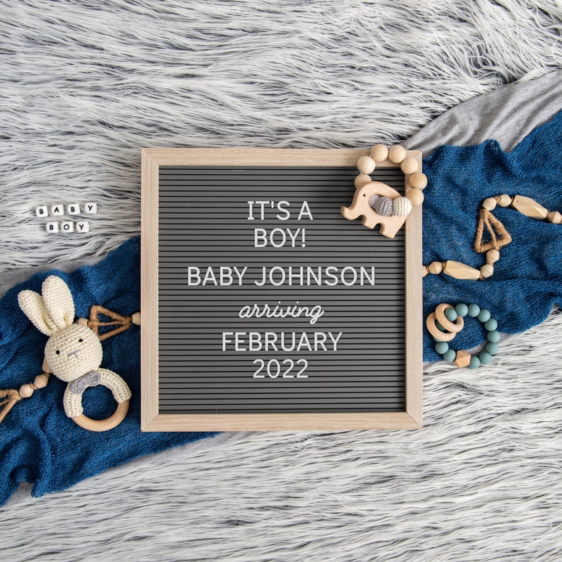 May include: A gray letter board with the text "It's a Boy! Baby Johnson arriving February 2022". The board is surrounded by a blue blanket, a wooden elephant toy, and a wooden bead rattle.