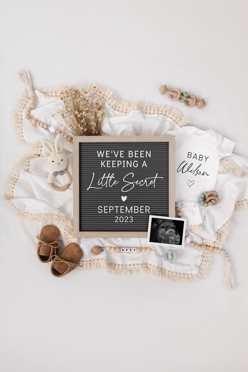 May include: A black letter board with the text "We've been keeping a little secret" and "September 2023".  A white onesie with the text "Baby Wilson" and a heart.  A wooden baby rattle, a pair of brown baby shoes, and a wooden bead garland with the word "Baby" spelled out.