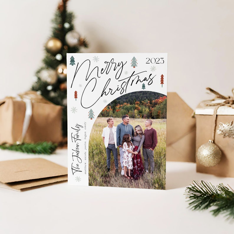 Pu&ograve; includere: Un biglietto di Natale con sfondo bianco e una foto di una famiglia di cinque persone in piedi in un campo. Il testo "Merry Christmas 2025" &egrave; scritto in corsivo nero con alberi stilizzati verdi e rossi e fiocchi di neve. Il cognome della famiglia "The Jackson Family" &egrave; scritto in nero con i nomi dei membri della famiglia elencati sotto.