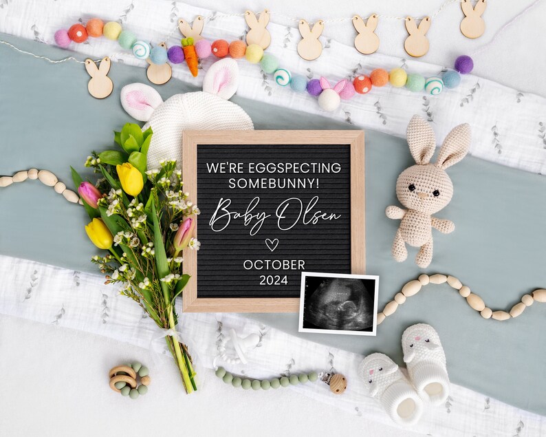 K&ouml;nnte beinhalten: Eine Buchstabentafel mit dem Text "We're eggspecting somebunny! Baby Olsen October 2024" umgeben von einem Strau&szlig; gelber und rosa Blumen, einem geh&auml;kelten Hasen und einem Baby-Ultraschallbild.
