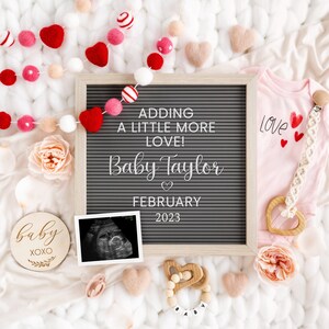 May include: A gray letter board with the text "Adding a little more love! Baby Taylor February 2023".  A pink onesie with the word "love" and hearts. A wooden baby rattle with the letters "baby" and a wooden ultrasound image.
