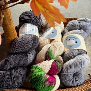 May include: A wicker basket holds several skeins of yarn in various colors. The yarn colors include dark gray, cream, and a vibrant mix of green and magenta. Each skein has a label that reads "YANI" and "HANDMADE".