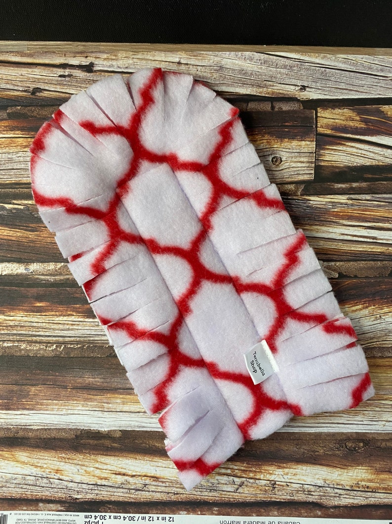 May include: White and red fleece dog toy with a fringe border. The toy has a geometric pattern and the text "Tenabella Shop" is printed on the toy.