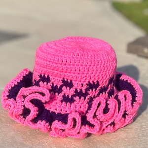 Peut inclure: Un chapeau de soleil en crochet rose avec un bord festonné et des accents violets.