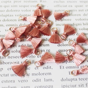 May include: A selection of small, dusky pink tassels with gold-coloured metal rings. These decorative tassels are likely used for jewellery making or craft projects. The tassels are arranged on a white surface with printed text.