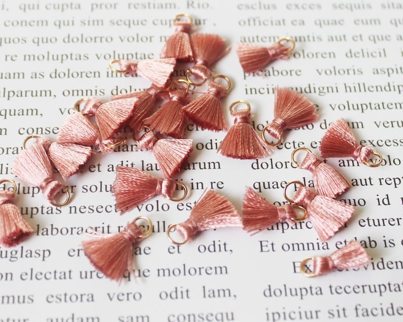 May include: A collection of small, dusty rose-colored tassels with gold-colored metal rings. These decorative tassels are likely used for jewelry making or craft projects. The tassels are arranged on a white surface with printed text.