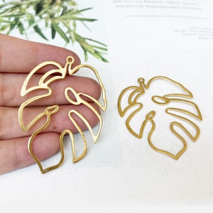 May include: Two gold-colored metal monstera leaf charms. The leaves are cut out and have a delicate, intricate design.