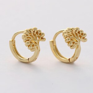 May include: A pair of gold-colored hoop earrings. Each earring features a detailed tree design on the front. The earrings have a hinged closure and are set against a white background.