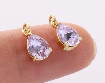 May include: Two gold-colored teardrop-shaped pendants with light purple gemstones. The gemstones are faceted and set in a gold-colored metal. The pendants are small and delicate, with a loop at the top for hanging.
