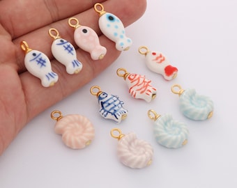 May include: A collection of ceramic charms in various shapes, including fish and seashells. The charms are painted in pastel colors like pink, blue, and white, with gold-colored metal loops for attaching to jewelry or other crafts.