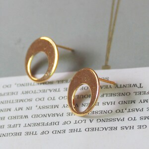 May include: Two gold-colored stud earrings. Each earring features a circular design with a crescent-shaped cutout. The earrings are displayed on a white surface with printed text, against a soft gray background.
