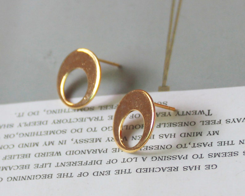 May include: Two gold-colored stud earrings. Each earring features a circular design with a crescent-shaped cutout. The earrings are displayed on a white surface with printed text, against a soft gray background.