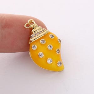 May include: A gold-colored charm in the shape of a seashell. The charm has a bright yellow enamel finish with clear crystal accents. The top of the charm is detailed with a band of small crystals.