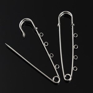 May include: Three silver safety pins with multiple loops on each pin. The pins are arranged in a triangular shape.