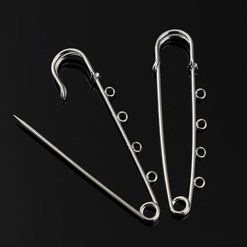 May include: Three silver safety pins with multiple loops on each pin. The pins are arranged in a triangular shape.
