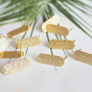 May include: A close-up of eight gold-toned oval-shaped earring blanks with textured surfaces. The earring blanks have a small hole at the top and a post on the back.