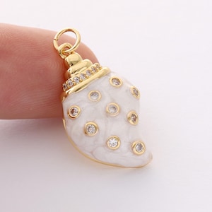 May include: A gold-colored seashell charm with a white enamel finish and clear crystal accents. The charm has a loop at the top for attaching to a necklace or bracelet. The seashell is a popular theme for jewelry.