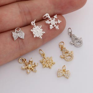 Puede incluir: Una colección de pequeños colgantes decorativos en tonos plateados y dorados. Los colgantes incluyen diseños de copos de nieve y muñecos de nieve, así como formas de lazo, todos adornados con pequeñas piedras brillantes. Estos colgantes son ideales para la creación de joyas.