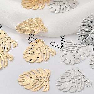 May include: A collection of gold and silver metal charms shaped like tropical leaves. The charms have a cutout design and are perfect for jewelry making or crafting.