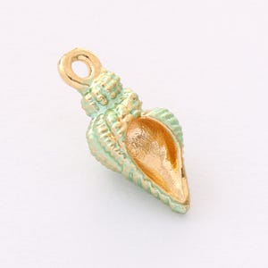 Puede incluir: Un pequeño dije en forma de concha con un lazo en la parte superior para colgar. El dije está pintado en color verde mar con detalles dorados y un interior dorado. La concha tiene una superficie texturizada y probablemente es de metal.