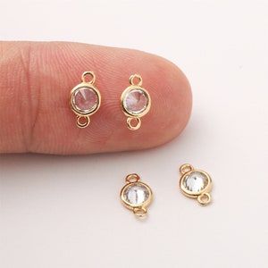 Puede incluir: Primer plano de varios conectores de bisel dorados con piedras preciosas claras, redondas y facetadas. Cada conector tiene un bucle en cada extremo. Los conectores están sobre un fondo claro.