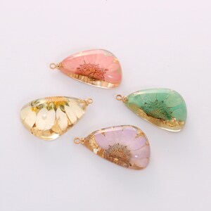 May include: Four teardrop-shaped resin pendants with dried flowers and gold-colored trim. The pendants contain pink, white, green, and purple flowers. Each pendant has a small loop for attaching to a chain or other jewelry component.