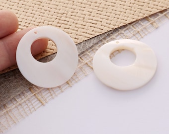 May include: Two white shell circle pendants with a hole in the center. The pendants are on a woven fabric background.