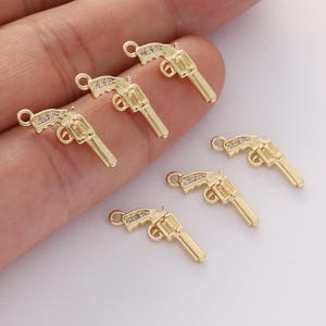 May include: Gold-colored gun charms with clear crystal accents. These small, detailed charms feature a classic revolver design, perfect for jewelry making or crafting projects. The charms are shown in a close-up view.