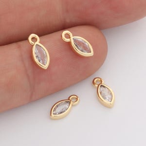 May include: Four small marquise-shaped charms featuring a clear, faceted crystal stone set in a polished gold-tone metal frame. Each charm includes a small circular jump ring at the top for attachment, showcasing a sleek and simple design.