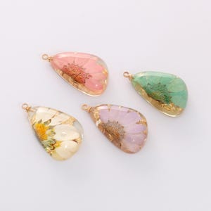 May include: Four teardrop-shaped pendants with dried flowers encased in clear resin. The pendants feature pink, green, purple, and white flowers with gold flakes. Each pendant has a small gold loop for hanging.