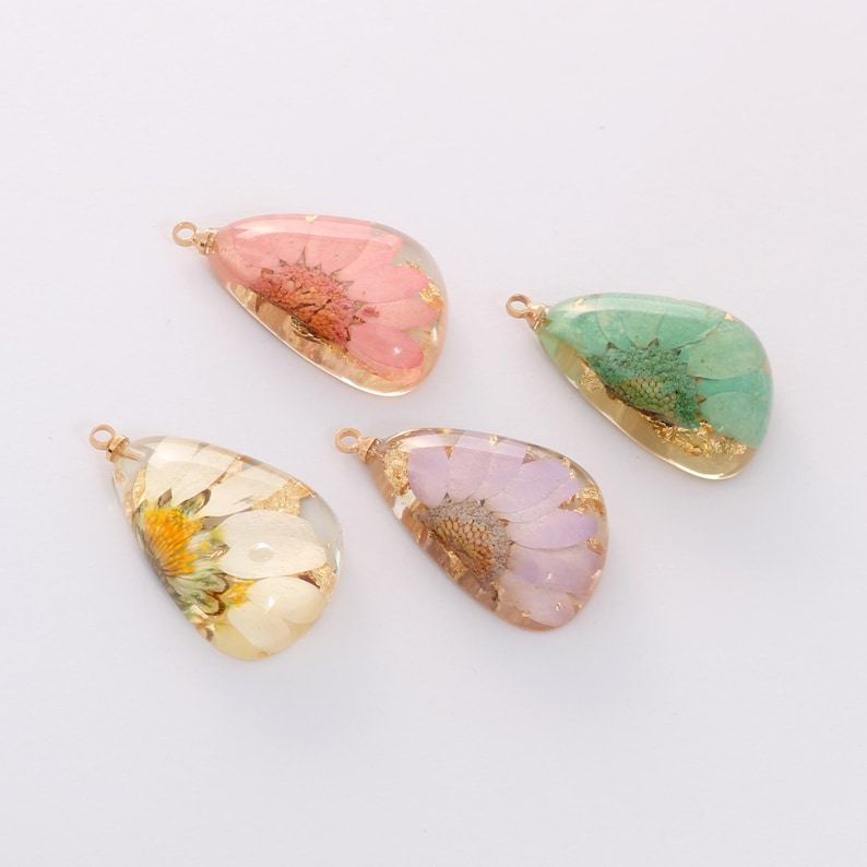May include: Four teardrop-shaped pendants with dried flowers encased in clear resin. The pendants feature pink, green, purple, and white flowers with gold flakes. Each pendant has a small gold loop for hanging.
