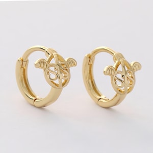 May include: A pair of gold hoop earrings with a delicate design featuring a circle with wings and the letters "SIS" inside.