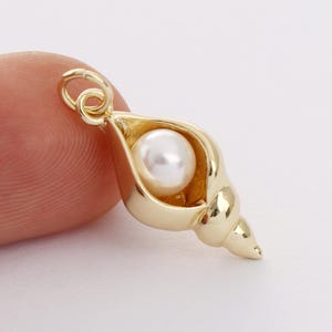 Pode incluir: Encanto de concha marítima em tom dourado com uma pérola branca dentro. O encanto tem um laço na parte superior para prender a um colar ou pulseira.