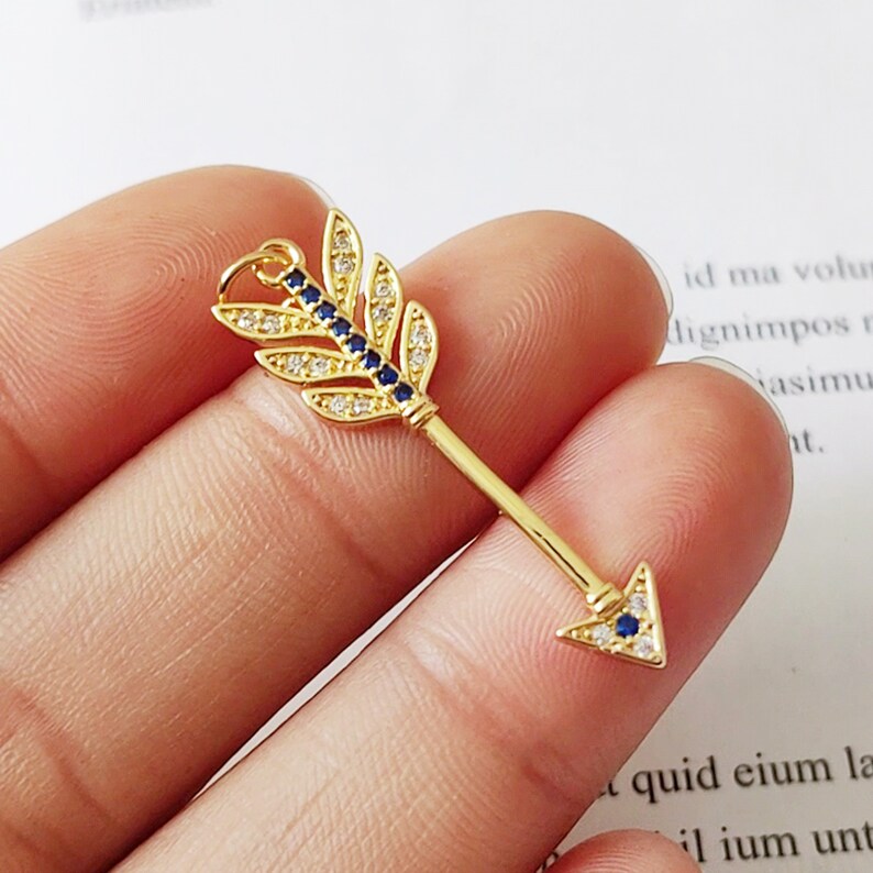 May include: Gold arrow charm with blue and white crystal accents. The arrow has a leaf-shaped design at the back and a pointed tip.