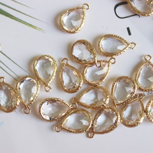 May include: A collection of clear glass teardrop-shaped pendants with gold-toned metal settings and loops. The pendants are scattered on a white surface.