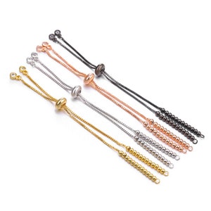 Op de afbeelding: Een set van vijf verstelbare armbandkoorden in verschillende metaalafwerkingen, waaronder goud, zilver, rosé goud en zwart. Elk koord heeft een kleine kraal aan het einde en een schuifkraal om de lengte aan te passen.
