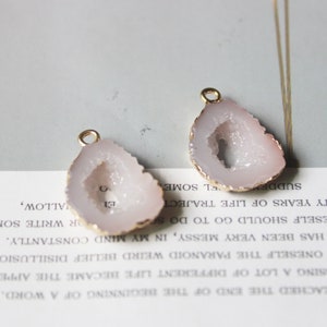 Puede incluir: Dos colgantes de ágata con borde dorado en un esquema de color rosa y blanco. El ágata es translúcida y tiene una formación de cristal natural.