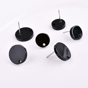 May include: Six black, circular stud earrings with silver posts are scattered on a white surface. Each earring has a small hole near the edge. The earrings are evenly spaced and appear to be made of a glossy material.