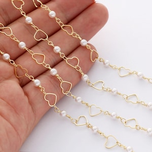 May include: Gold heart-shaped chain with white pearl beads. The chain is shown on a hand, with multiple strands of the chain.