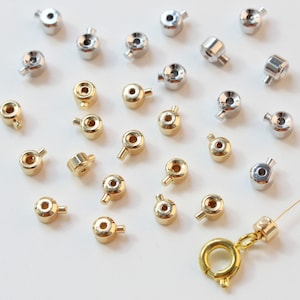 May include: A collection of gold and silver metal bead caps with a small loop on the top. The bead caps are arranged on a white surface.
