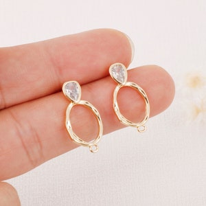 May include: A pair of gold-tone earrings with a teardrop-shaped clear crystal at the top of each earring. The earrings have a textured, hammered look and a circular hoop.