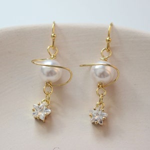 May include: Gold earrings with a pearl and a star-shaped crystal. The pearl is suspended from a gold wire that is twisted into a spiral shape.